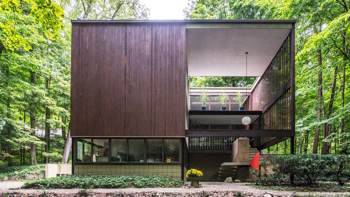 A geometric home with a solid side with wood siding and an ‘open’ side with a lofted living space. The home is surrounded by woods.